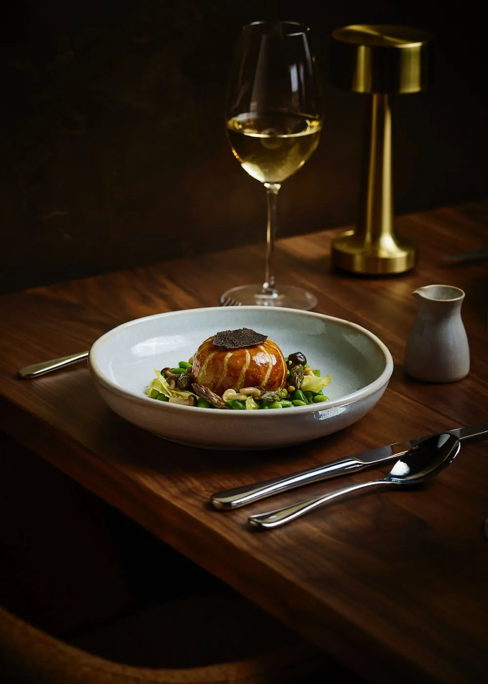 Gourmet plated dish with a glazed, round pastry topped with truffle slices, surrounded by green vegetables and mushrooms, accompanied by a glass of white wine on a wooden table.