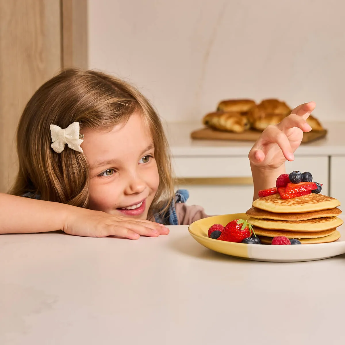 Smiling young girl with a hair bow reaching for a stack of pancakes topped with berries on a plate.