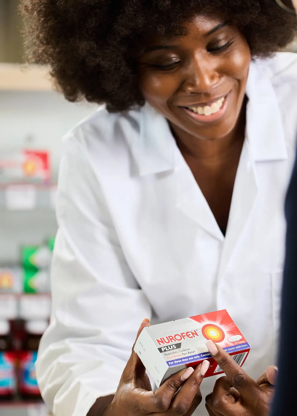 Smiling pharmacist holding and showing a box of Nurofen Plus pain relief tablets to a customer.