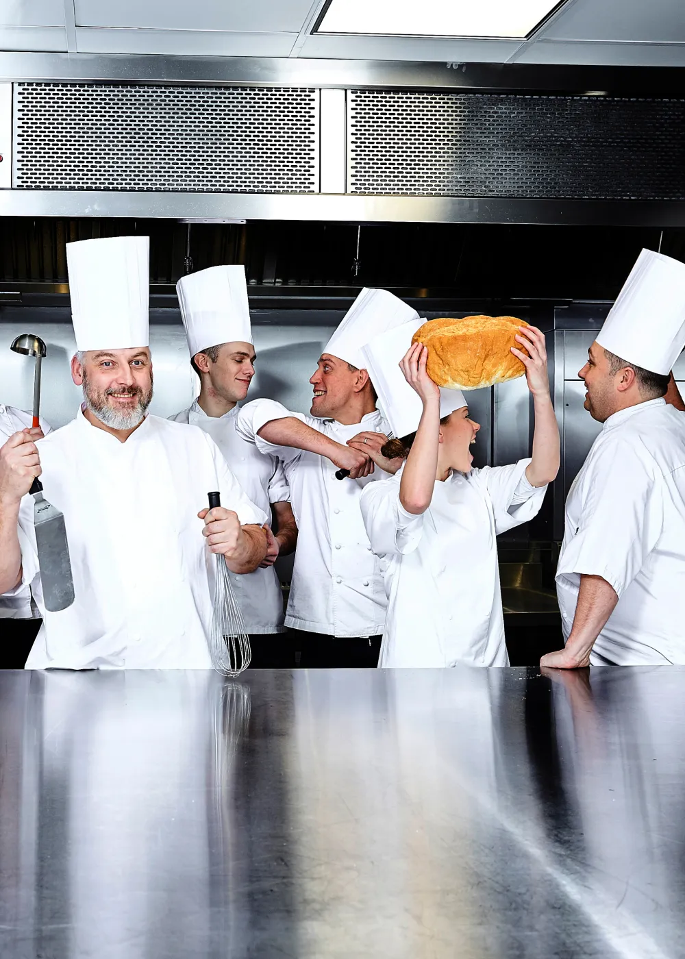 Group of five chefs in white uniforms and tall hats in a professional kitchen, some holding kitchen tools and one holding a loaf of bread above their head.