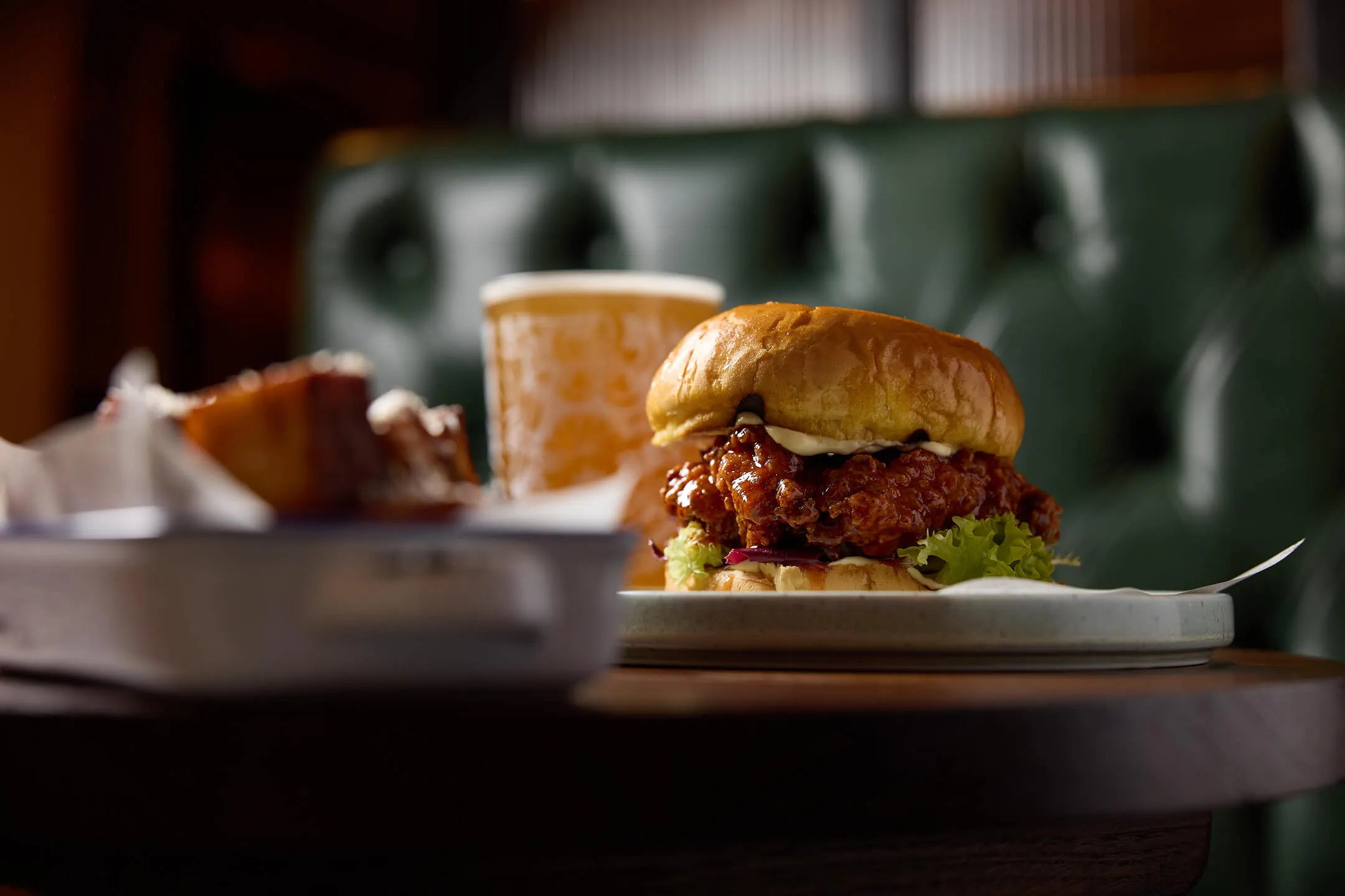 Close-up of a crispy fried chicken sandwich with lettuce and sauce on a plate, with a blurred drink and dish in the background.