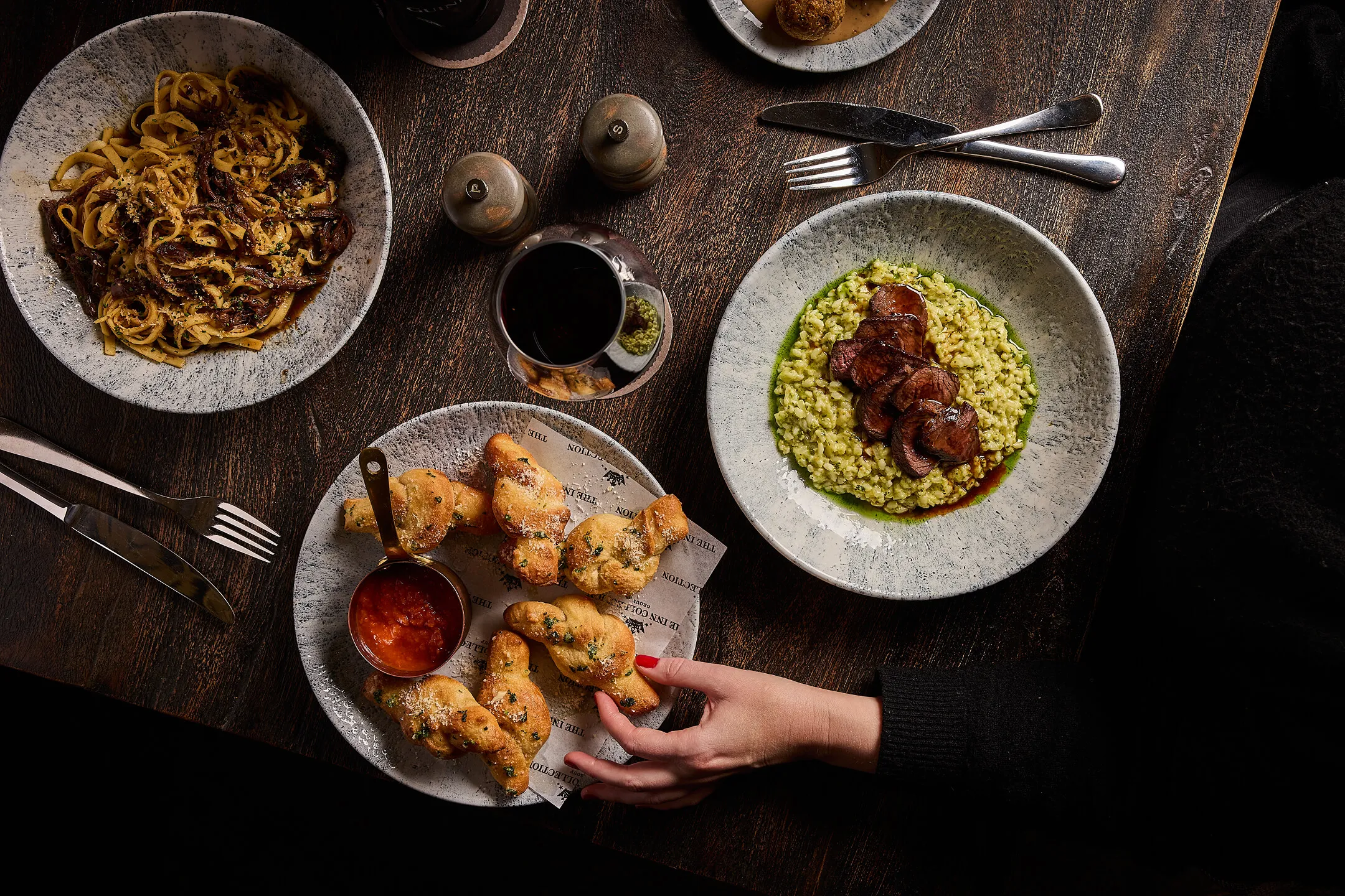 Table with plates of mushroom pasta, risotto topped with sliced meat, fried dough knots with marinara sauce, a glass of red wine, and a hand reaching for the bread.