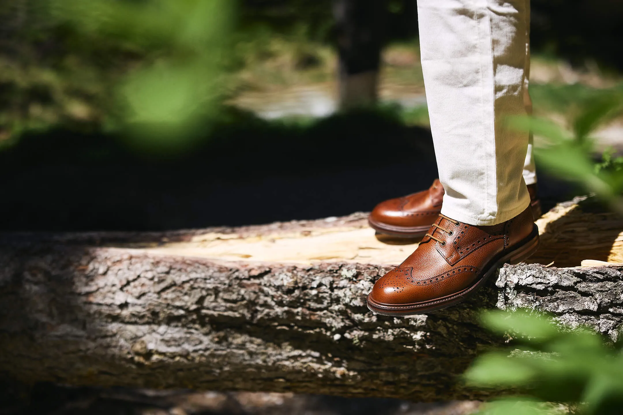 Brown leather brogue shoes worn with light-colored pants standing on a large tree log outdoors.
