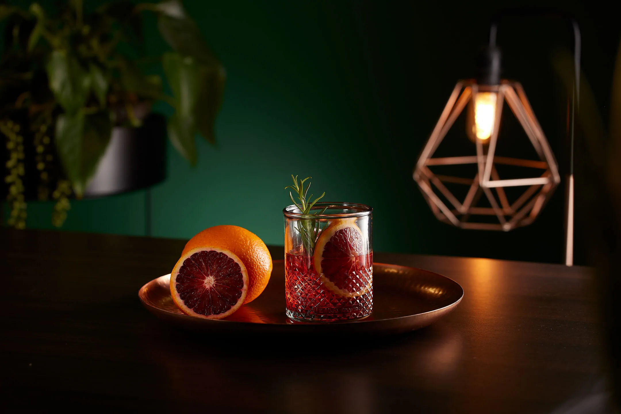 Glass with a red cocktail garnished with a slice of blood orange on a copper plate beside a halved blood orange, with a geometric lamp in the background.