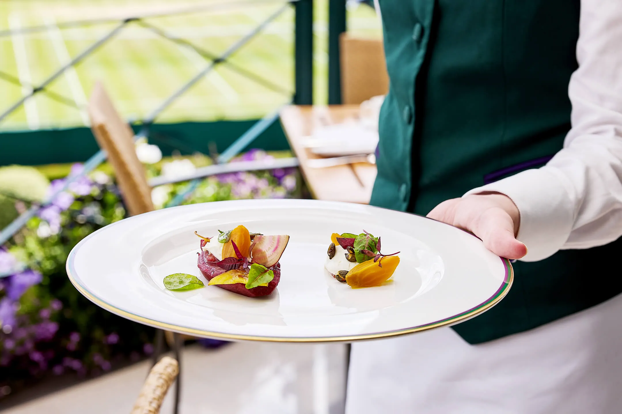 Waiter holding a white plate with two small gourmet vegetable dishes garnished with leaves and seeds with Wimbledon in the background.