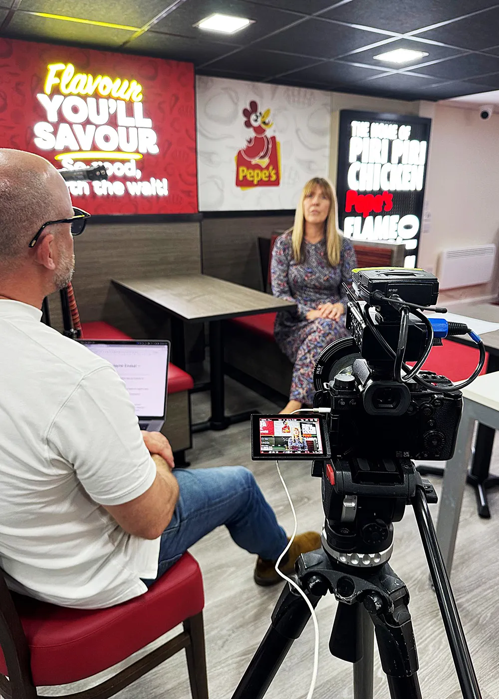 Woman sitting on a bench inside Pepe's restaurant being interviewed on camera by a man with a laptop.