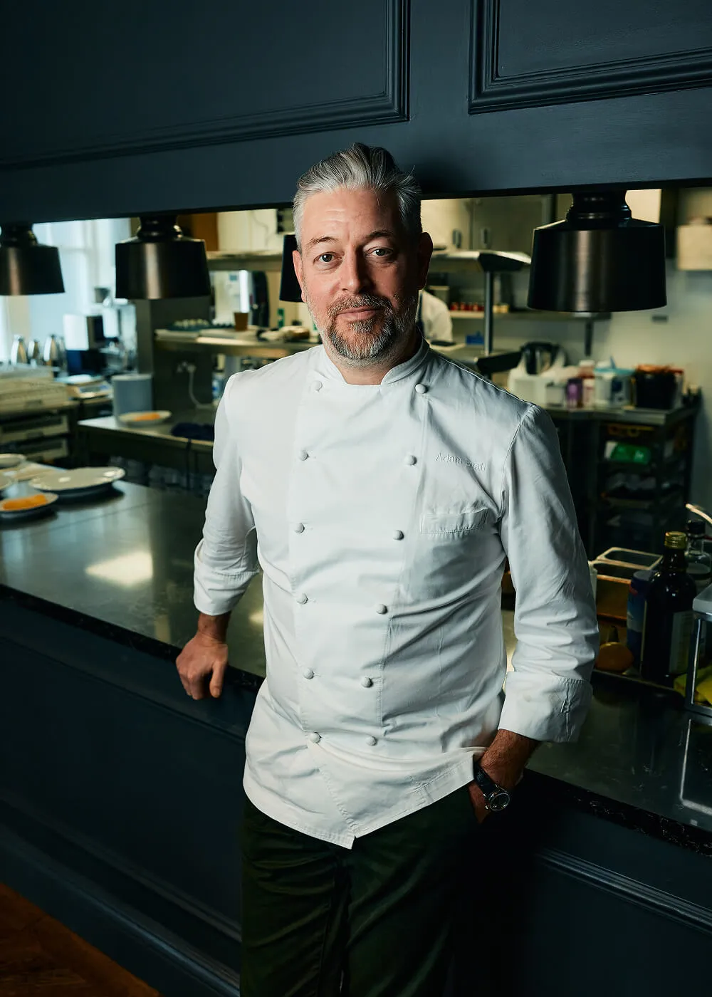 Male chef with grey hair and beard wearing a white double-breasted chef coat, standing in a professional kitchen.