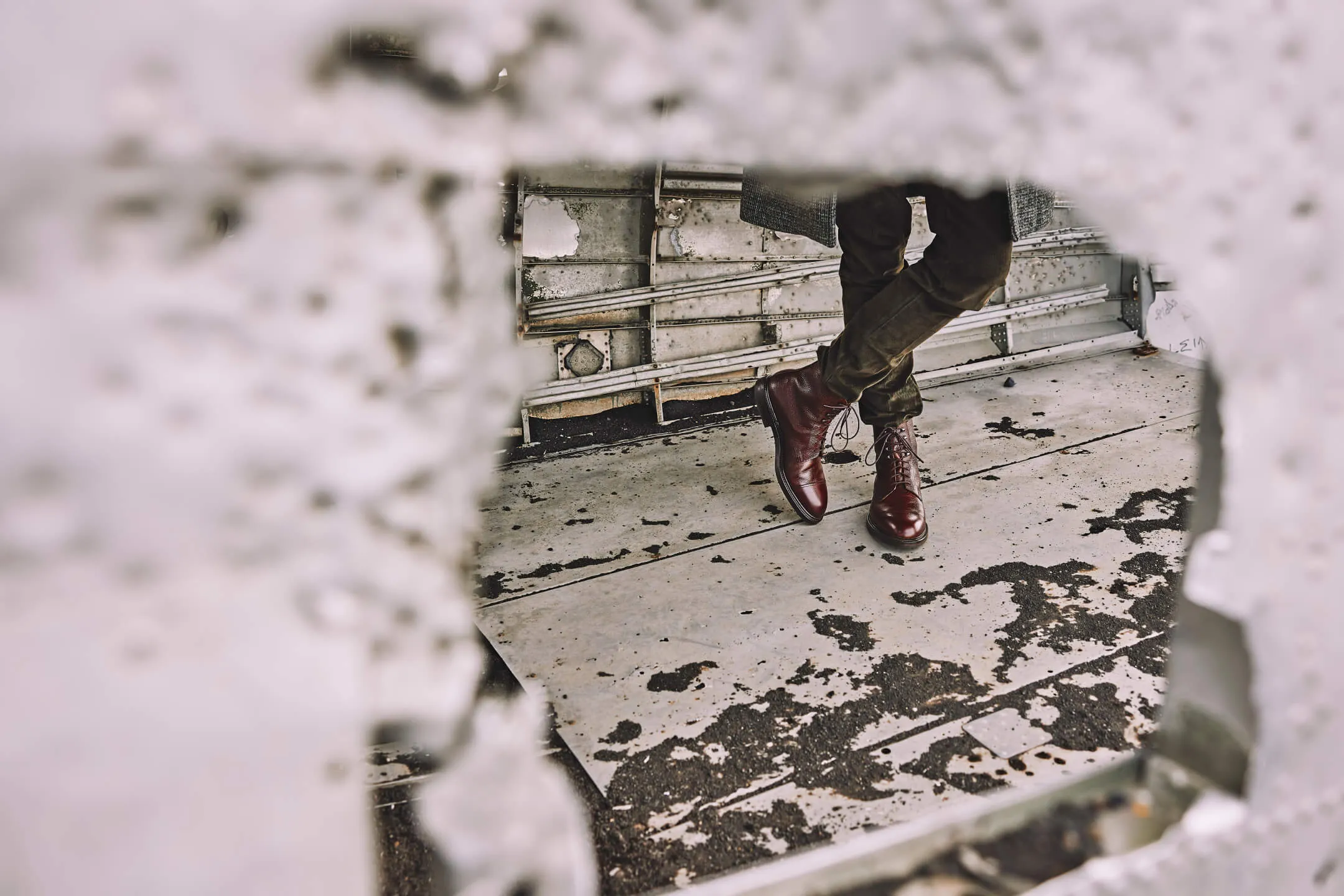 Person wearing brown leather lace-up boots standing on a weathered concrete floor, viewed through a blurred irregular hole.