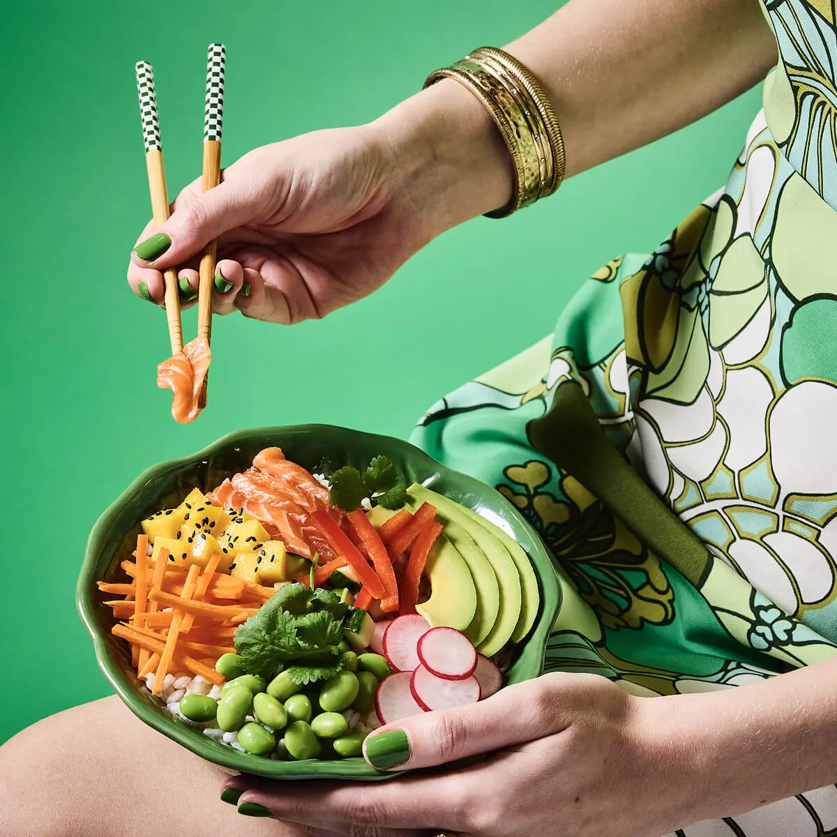 Person holding a green bowl of poke bowl with sliced avocado, radish, edamame, carrots, mango, red pepper, salmon, and cilantro, picking up a piece of salmon with chopsticks.
