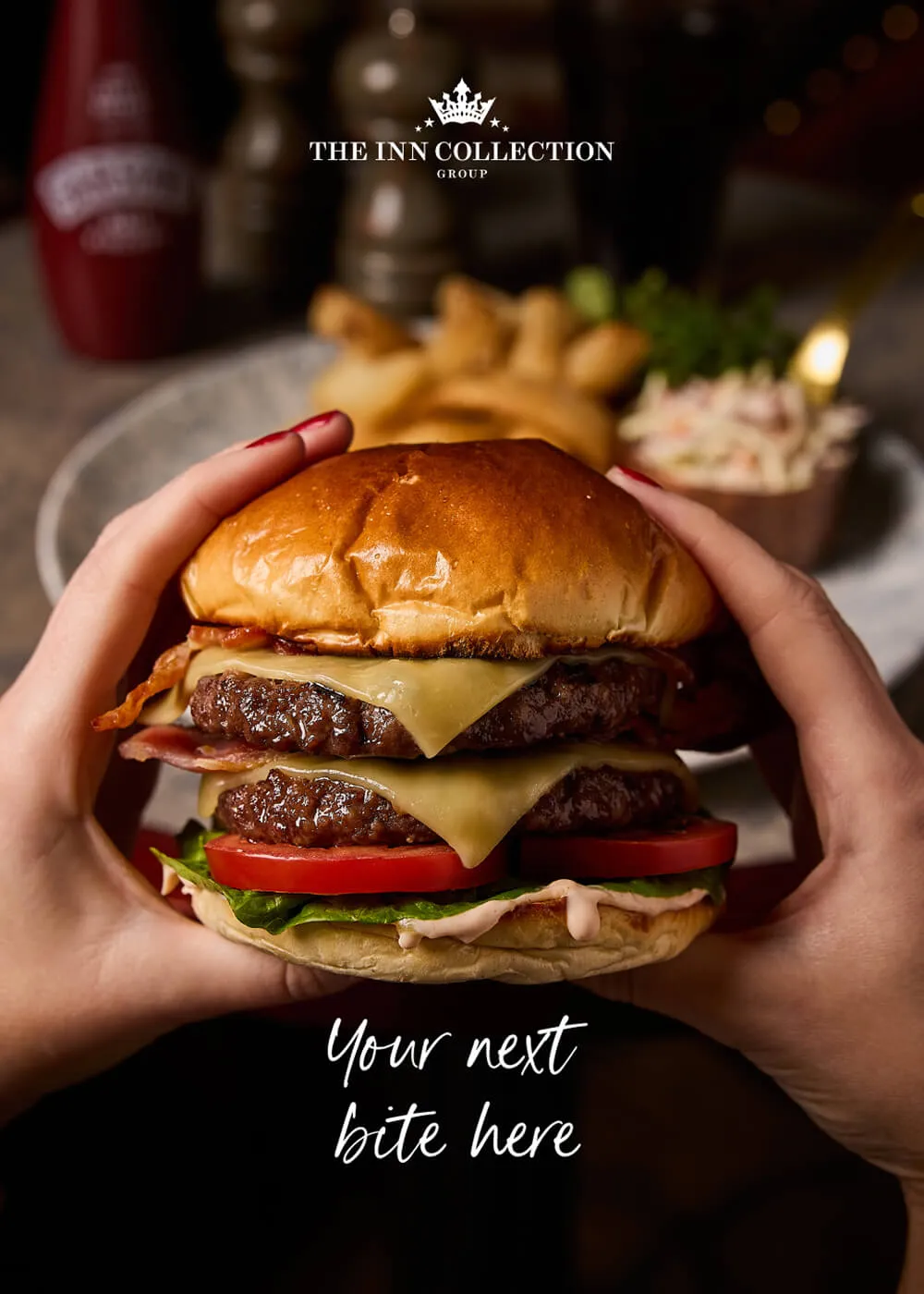 Hands holding a double cheeseburger with lettuce, tomato, bacon, and melted cheese with a plate of fries and coleslaw blurred in the background.