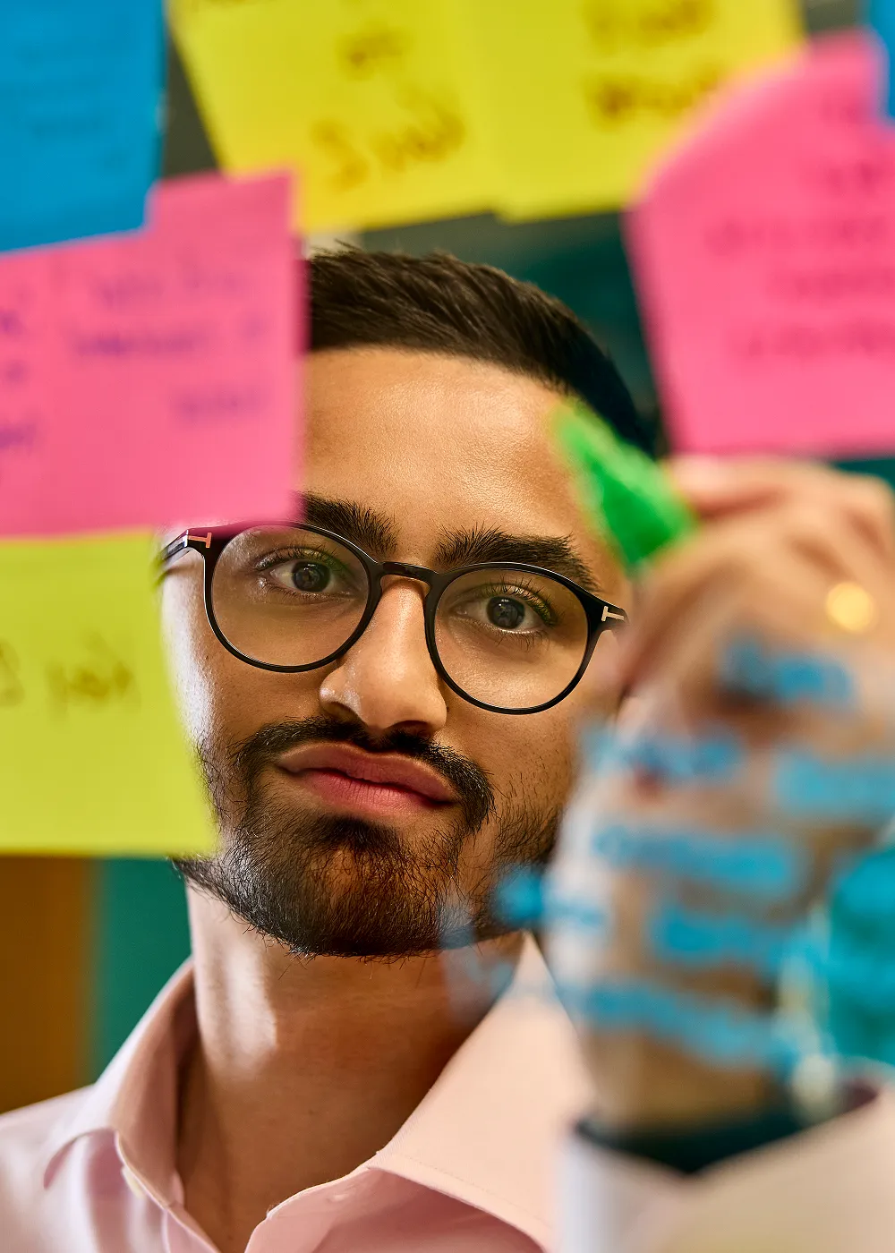 Young man wearing glasses writing on a glass wall surrounded by colourful sticky notes.