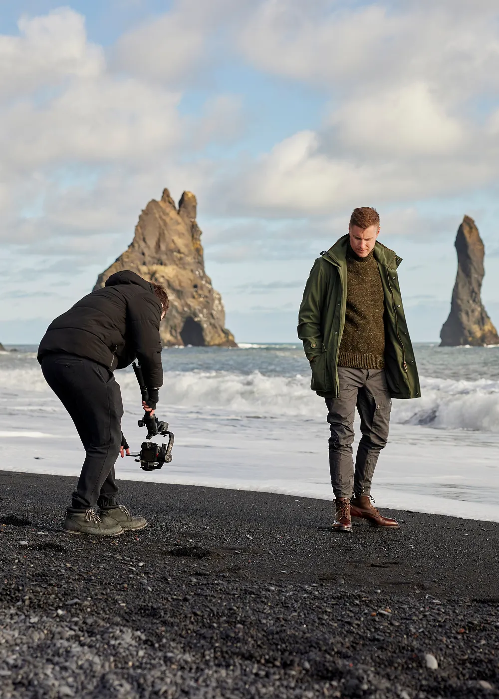 Man in green jacket and brown boots walking on a black sand beach while another person films him with a camera stabiliser, with large rock formations in the ocean in the background.