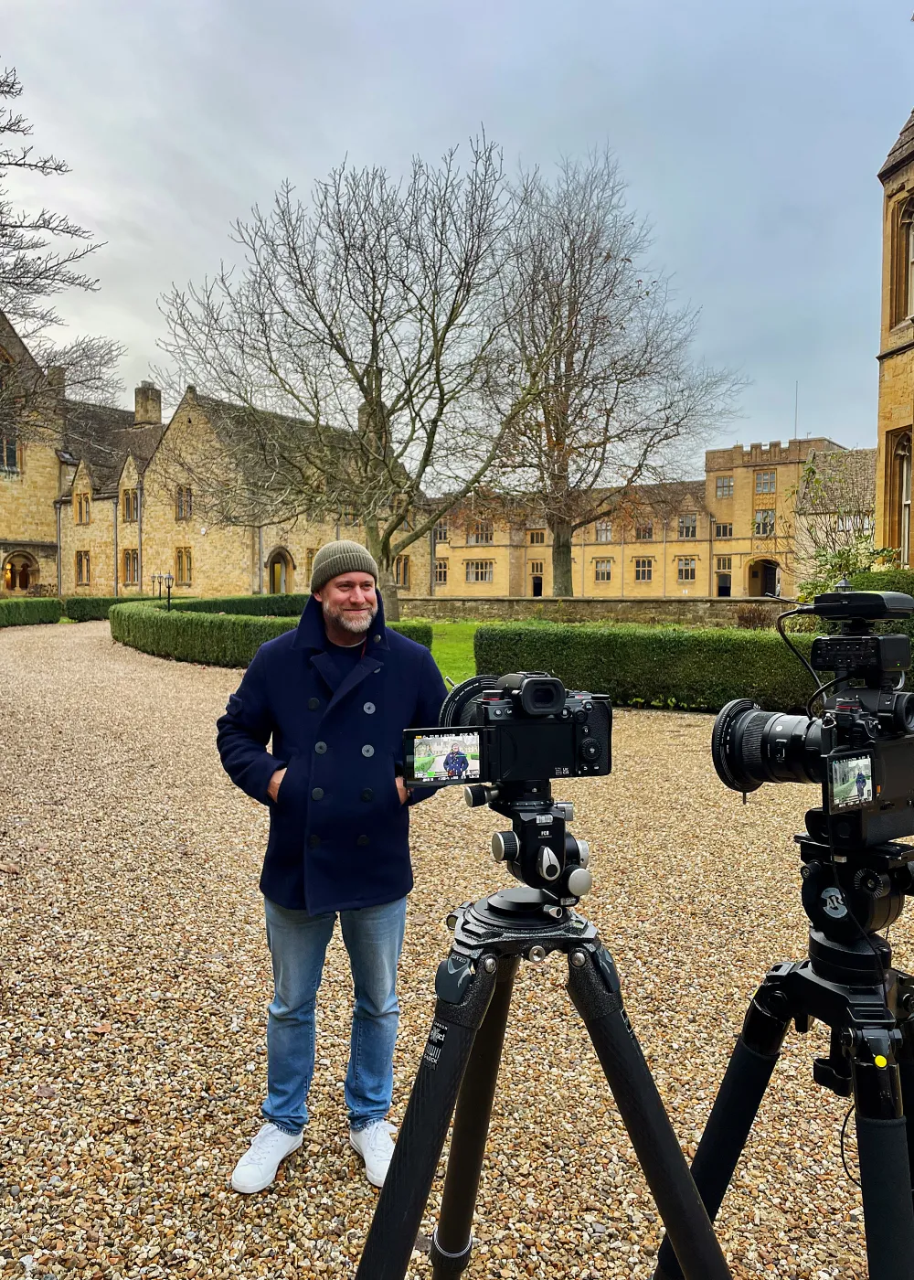 Man wearing a navy coat and green beanie standing on gravel beside two cameras on tripods in front of historic yellow stone buildings and leafless trees.