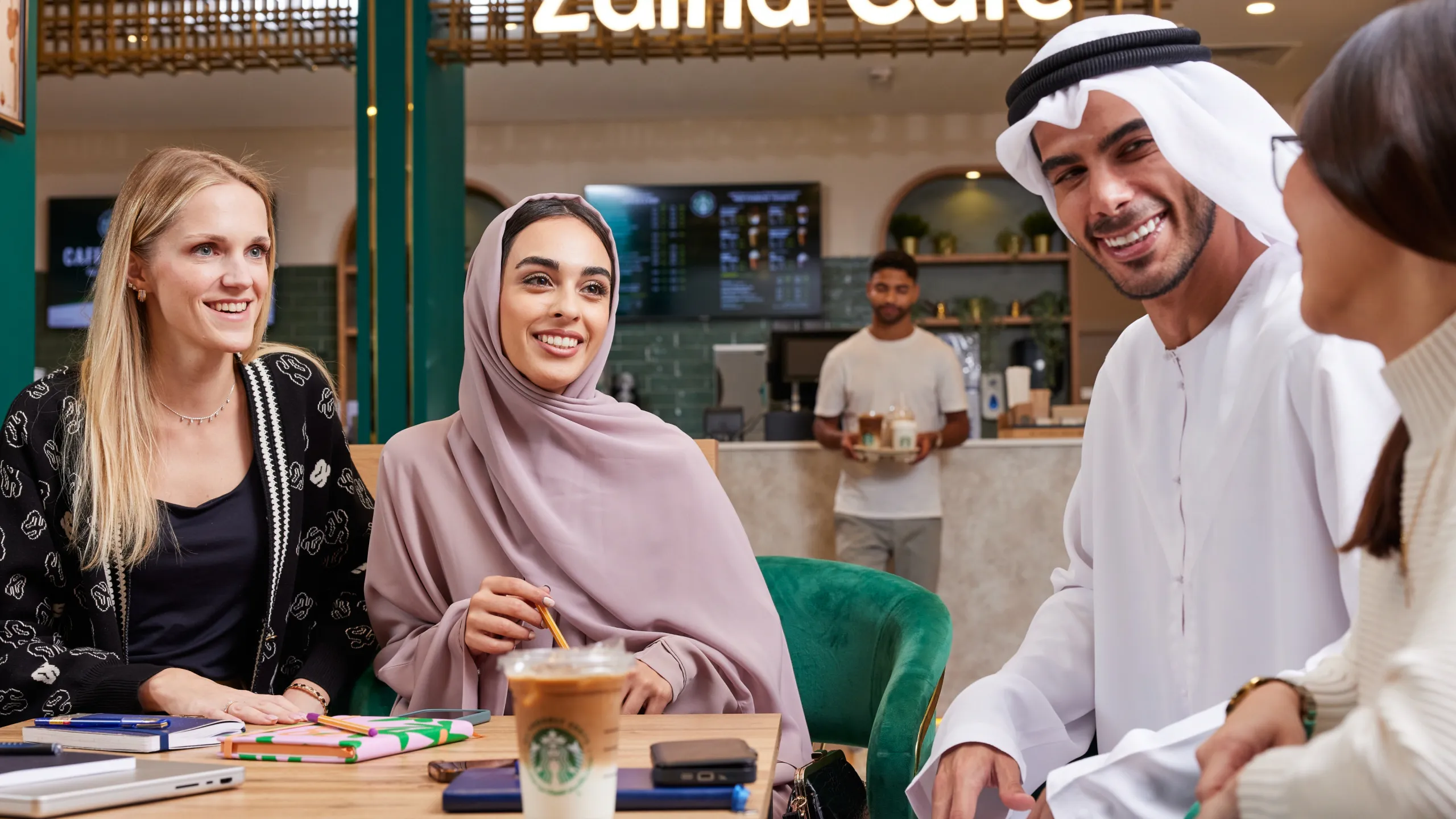 Four friends smiling and chatting at a cafe table with notebooks and drinks, a barista carrying coffee in the background.