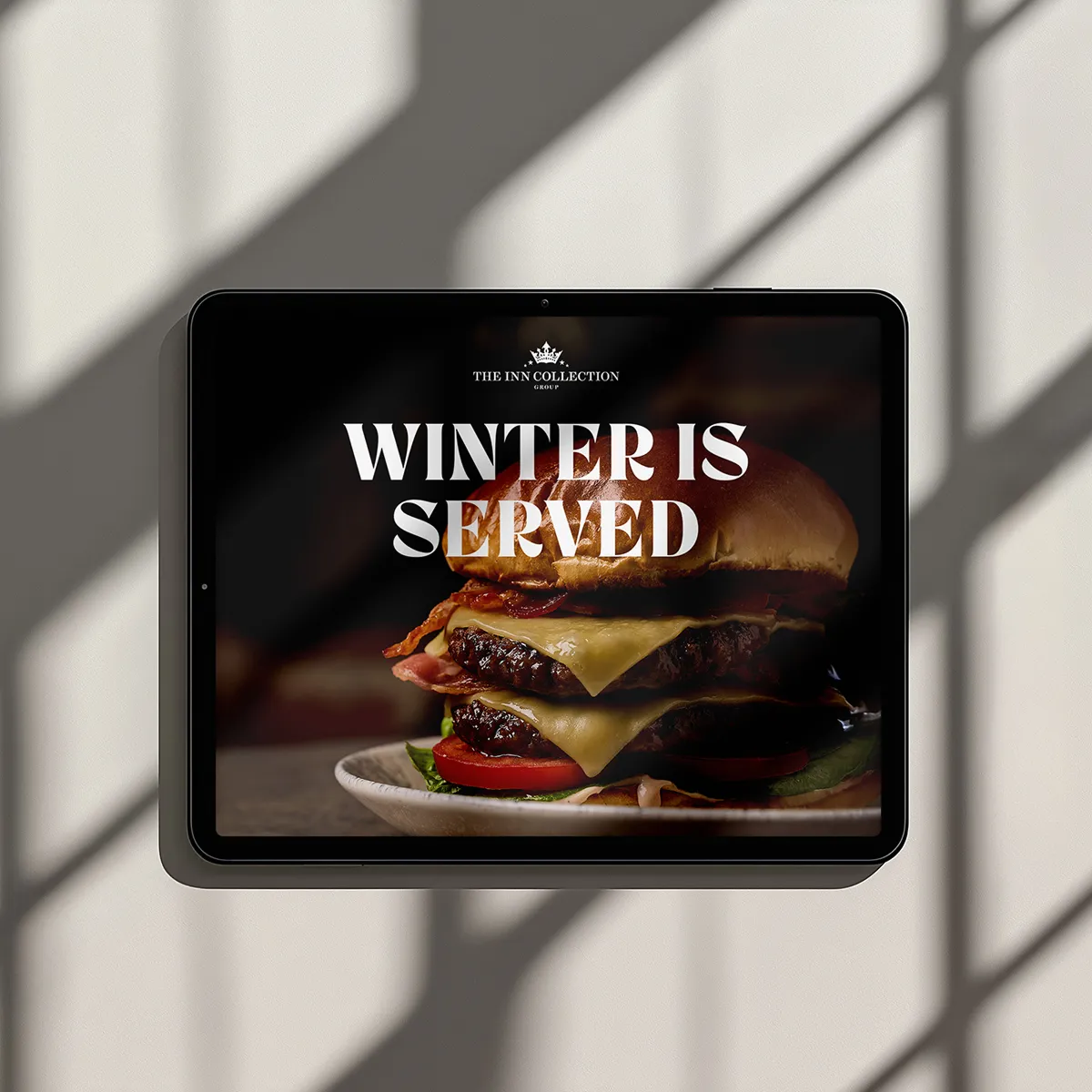 Tablet displaying a double cheeseburger with bacon and vegetables, overlayed with text 'Winter is Served' from The Inn Collection.