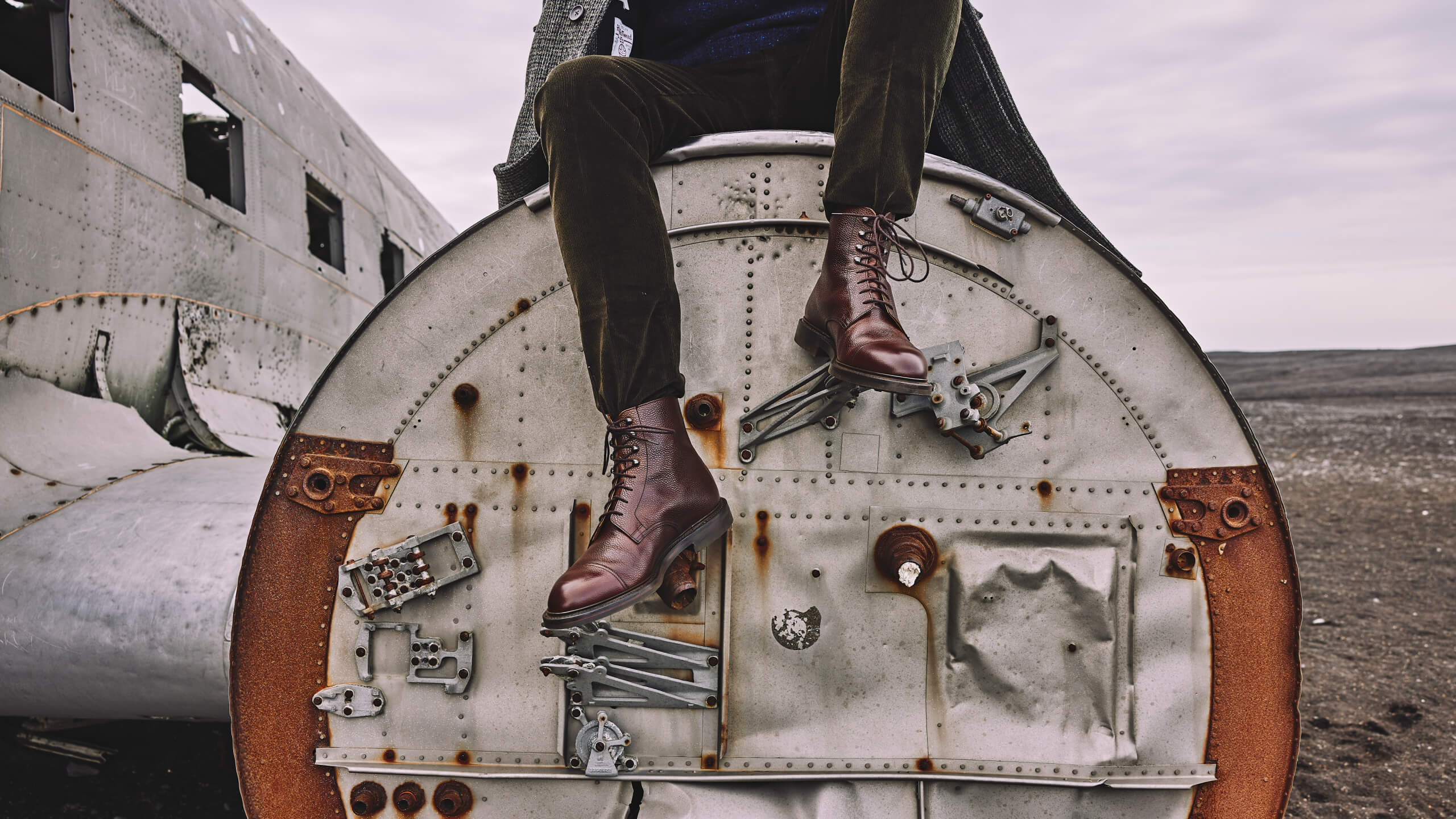 Person wearing Crockett & Jones brown leather boots and dark pants sitting on the rusted nose of an abandoned plane in a barren landscape.