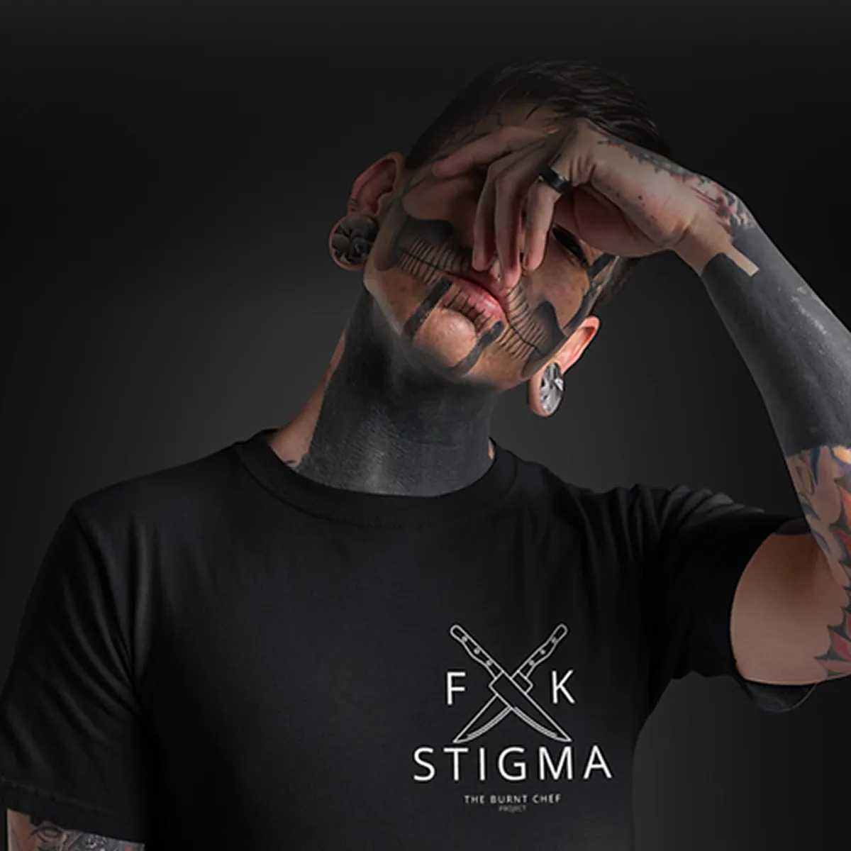 Person with extensive black tattoos on face, neck, and arm wearing black t-shirt with crossed knives and text 'F K STIGMA The Burnt Chef Project'.
