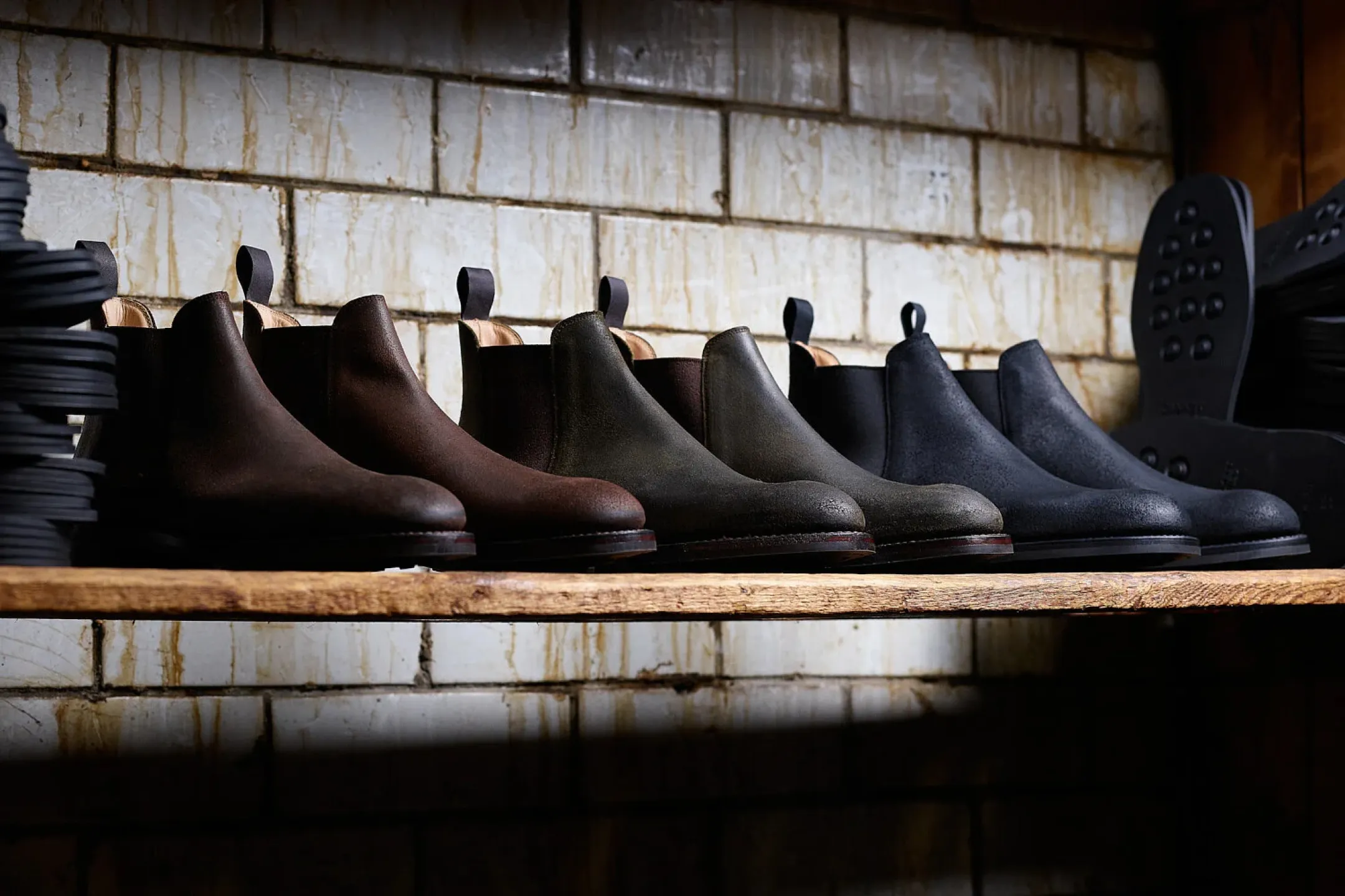 Row of six leather Chelsea boots in varying shades of brown, green, and black on a wooden shelf against a stained white brick wall.