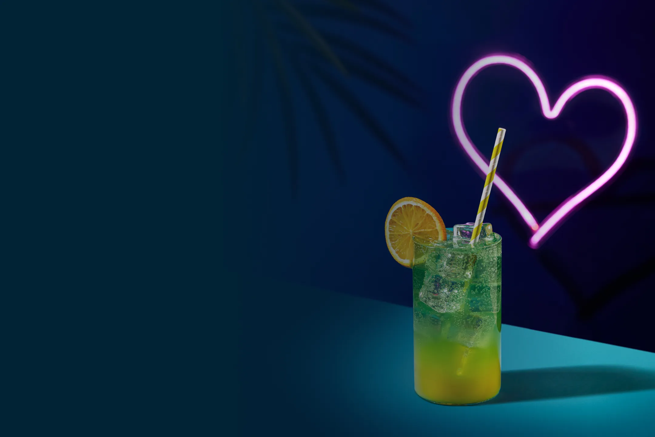 Tall glass of layered green and yellow tropical cocktail with ice, a lemon slice, and striped straw, set on a blue surface with a glowing pink neon heart in the background.