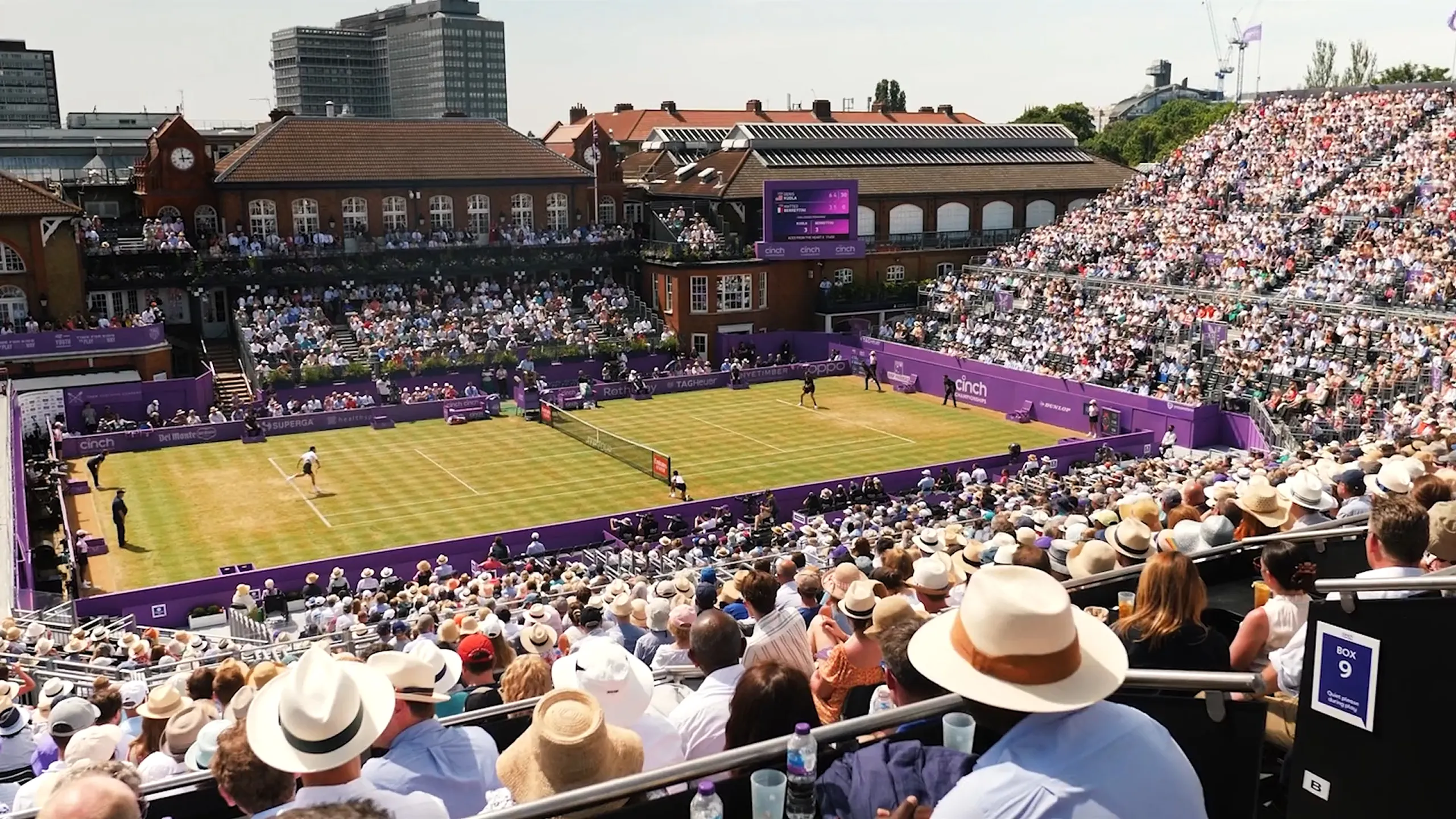 Outdoor grass tennis court at Wimbledon with a large crowd watching a match on a sunny day, surrounded by historic buildings and purple banners.