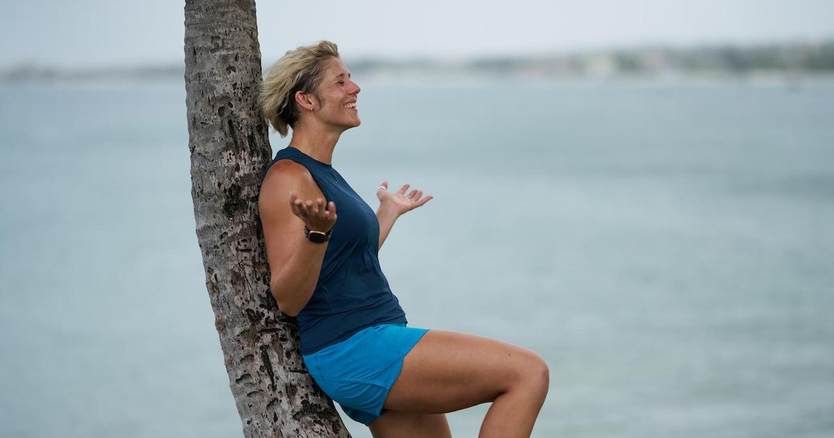 Ruth Mac leaning against a palm tree, smiling and holding her arms out to her sides.