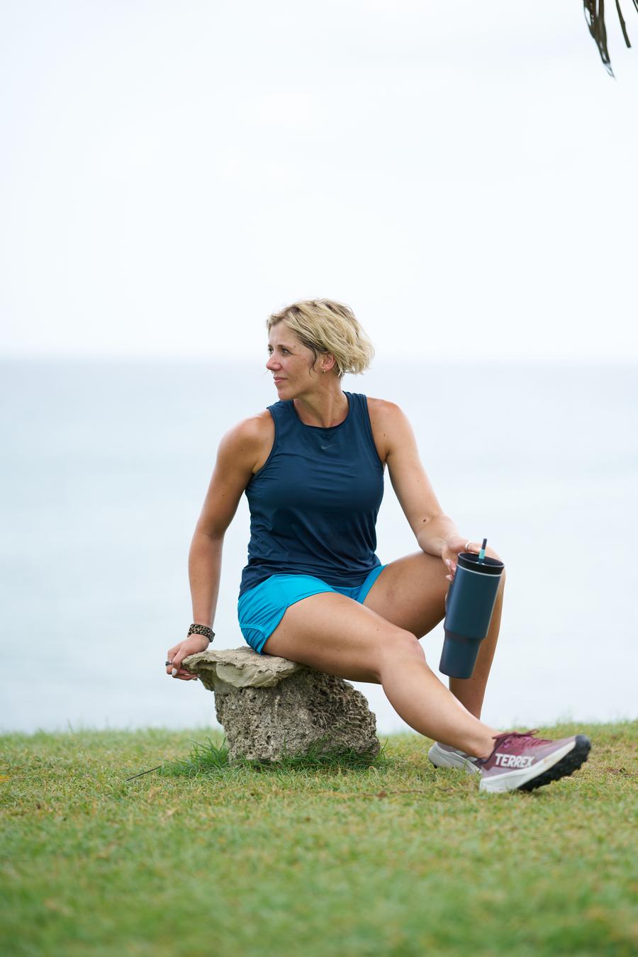 Ruth Mac sitting on a tree stump, holding a drinking flask, and looking over her shoulder with the sea in the background.