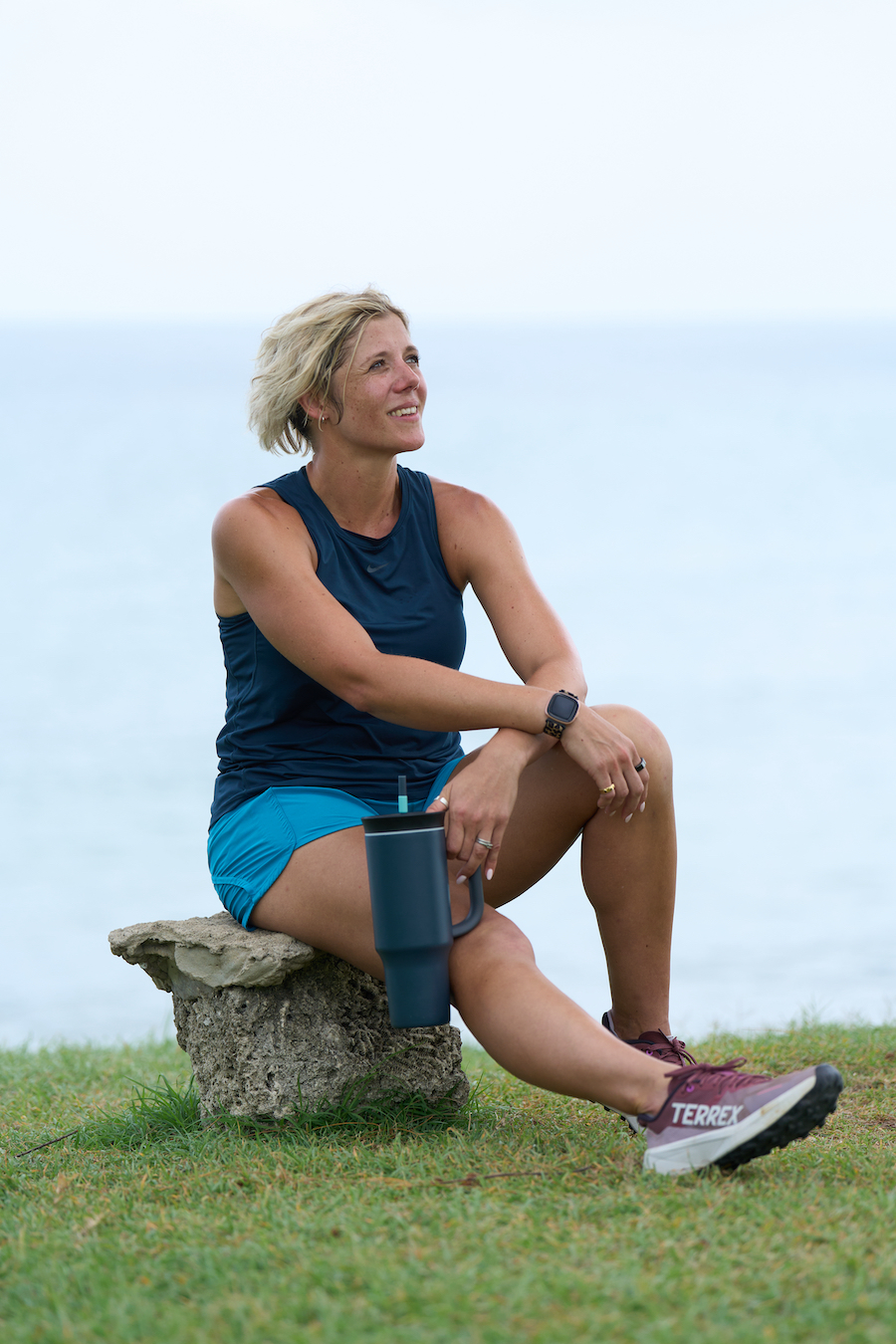 Ruth Mac sitting on a tree stump holding a drink, with the sea in the background.