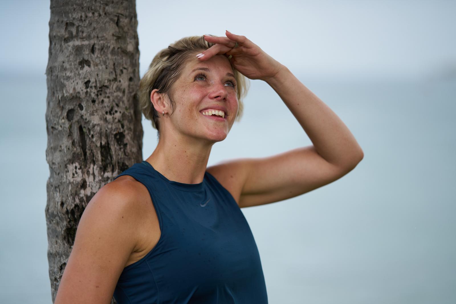 Ruth Mac leaning against a palm tree and looking up at the sky, with the sea in the background.
