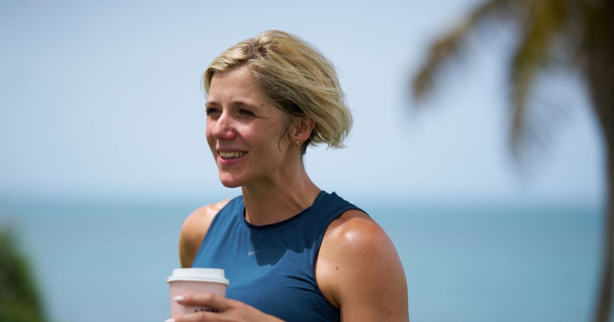 Ruth Mac holding a hot drink in a cardboard cup, with the sea in the background.