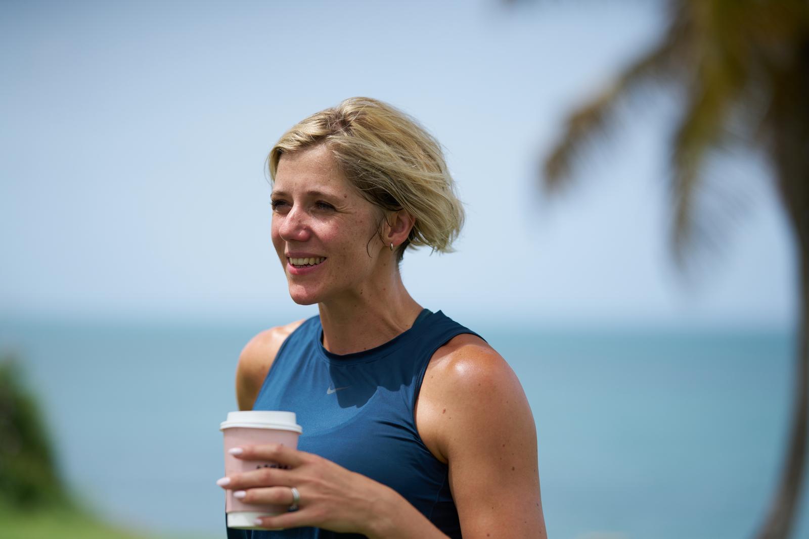 Ruth Mac holding a hot drink in a cardboard cup, with the sea in the background.