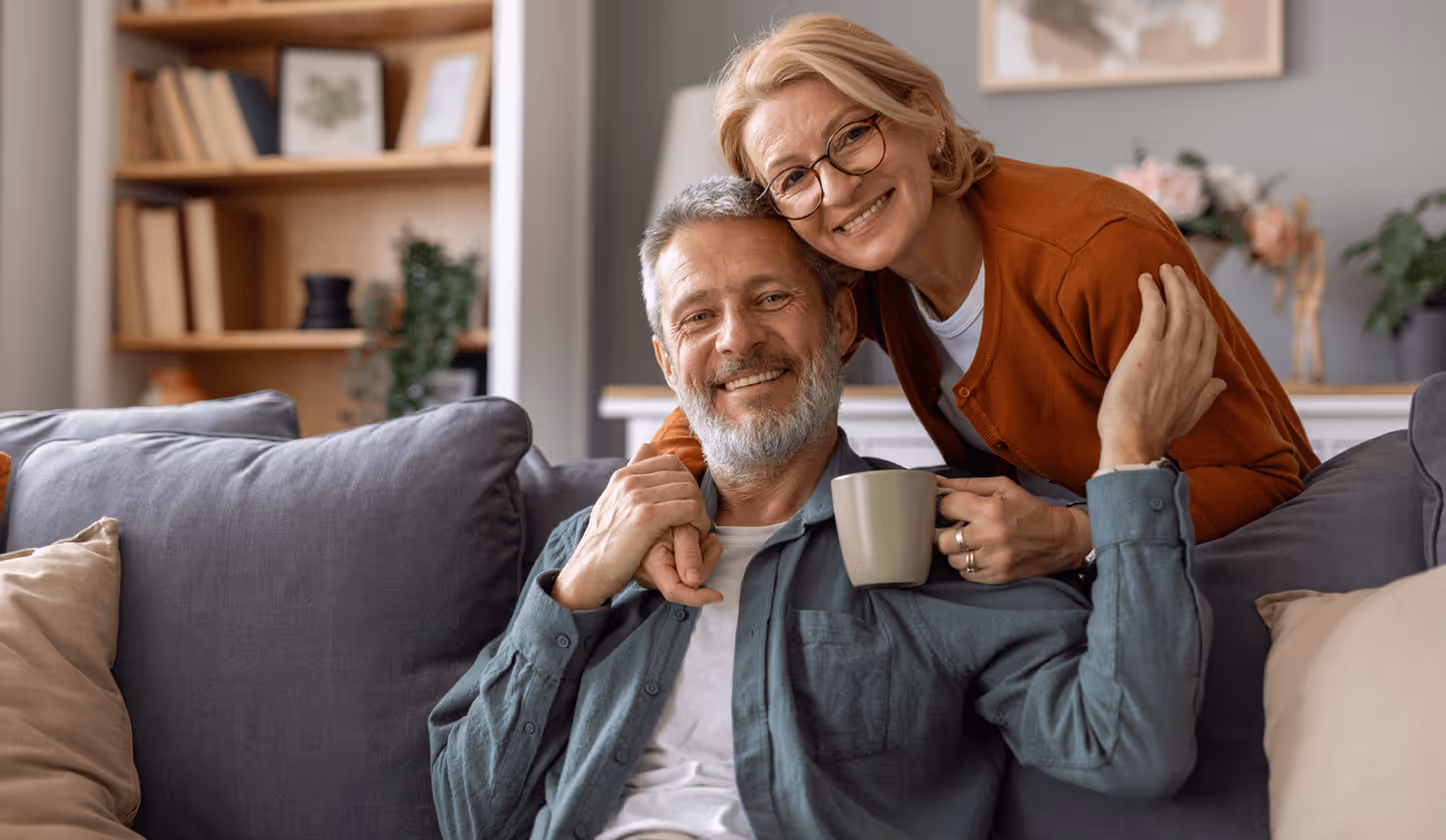 Älteres Paar lächelnd auf Sofa, Mann hält Tasse, Frau lehnt sich an ihn, gemütliche Wohnzimmeratmosphäre.
