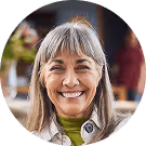 Ältere Frau mit grauem Haar, lächelt herzlich, trägt grüne Kleidung. Verschwommener Hintergrund mit warmen Tönen.