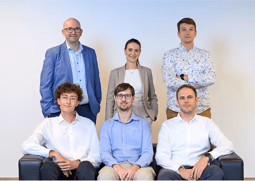 Sechs Personen lächeln professionell im Büro, drei sitzend, drei stehend, formelle Kleidung, neutraler Hintergrund.