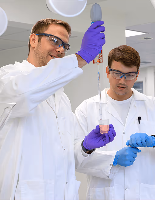 Zwei Wissenschaftler in weißen Kitteln führen im Labor eine Messung mit einer Pipette und farbiger Flüssigkeit durch.