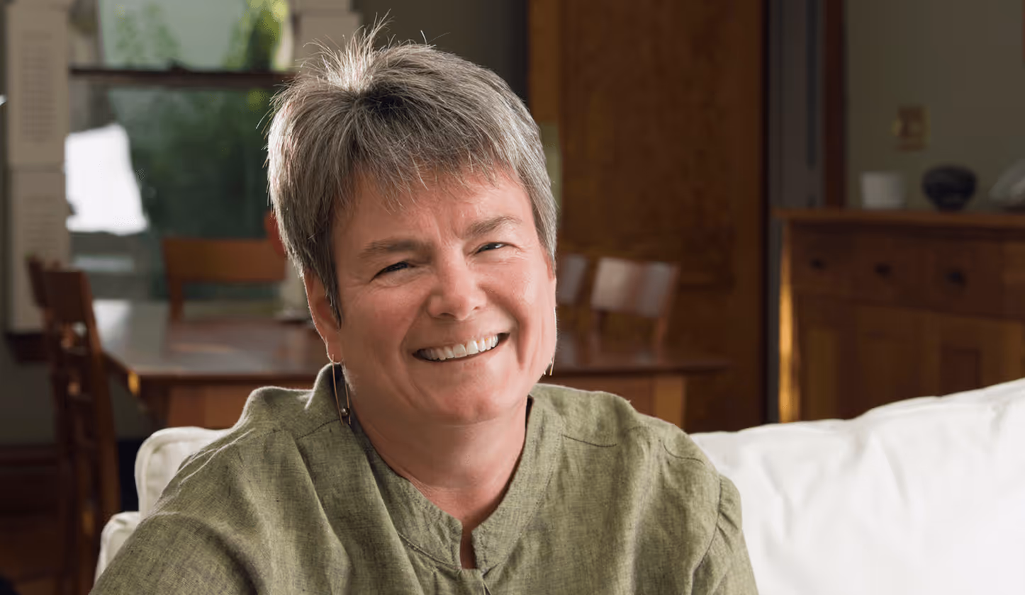 Lächelnde Person auf einem Sofa im Wohnzimmer, Holzschränke im Hintergrund.