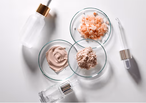 Glas-Schalen mit rosa Salzkristallen, Tonerde und Cremes, umgeben von Pipettenflaschen auf weißem Hintergrund.