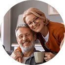 Älteres Paar lächelnd auf Sofa, sie umarmt ihn, er hält eine Tasse. Gemütliche Wohnzimmeratmosphäre.