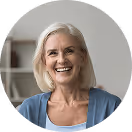 Lächelnde ältere Frau mit grauem Haar in blauem Pullover, sitzt vor unscharfem Raumhintergrund.