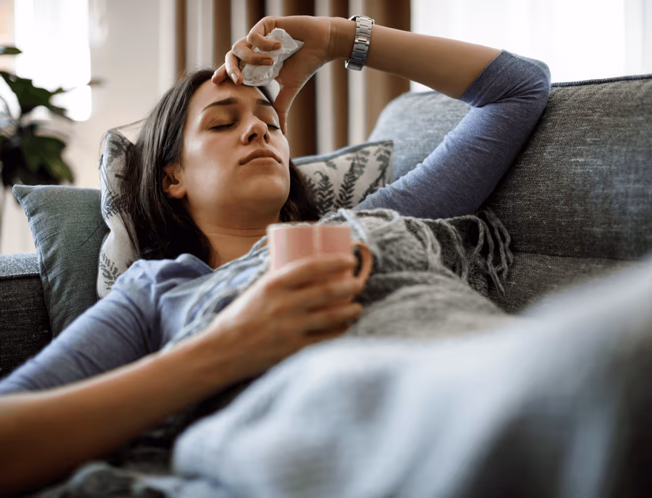 Frau mit geschlossenen Augen liegt krank auf Sofa, hält Teetasse und tupft Stirn mit Taschentuch.