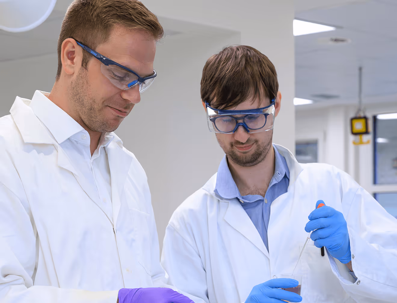 Zwei Männer im Labor mit Schutzbrillen und Handschuhen arbeiten konzentriert mit Pipette.