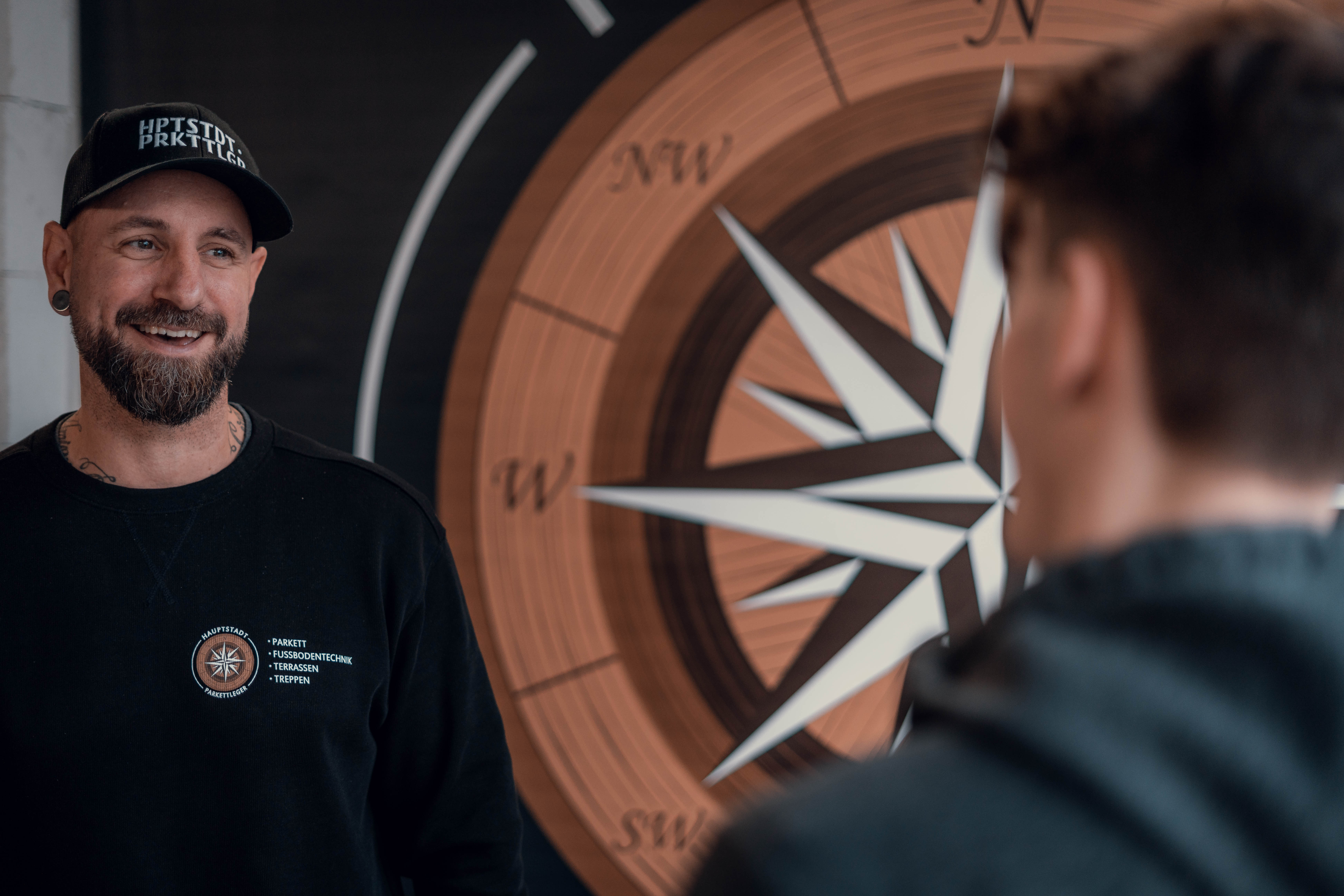 Alexander Kempkes im Gespräch mit einem Kunden oder Kollegen vor dem Logo von Hauptstadt Parkettleger im Showroom