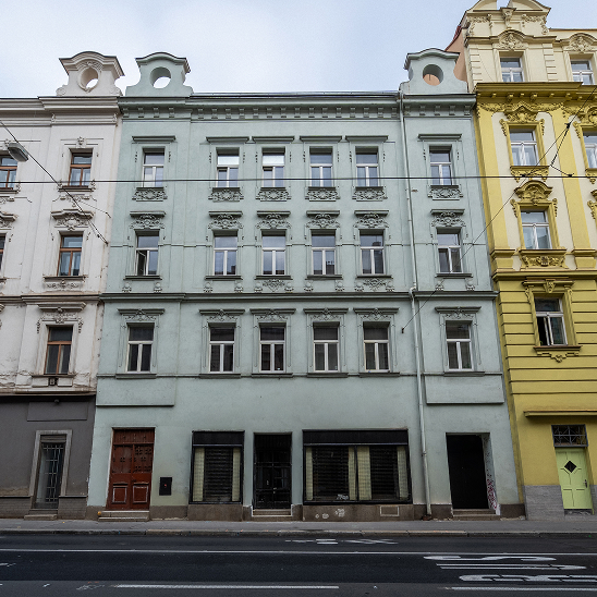 Fasáda staré budovy v šedozelené barvě se zdobenými okny mezi dvěma barevnými budovami na městské ulici.