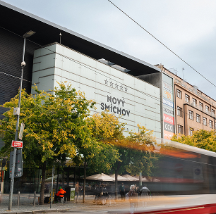 Fasáda obchodního centra Nový Smíchov za denního světla s jedoucím tramvajovým vagónem a stromy s žlutým listím v popředí.
