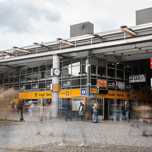 Lidé čekající před vchodem do budovy veřejné dopravy se žlutými orientačními tabulemi a obchodem Pizza Colosseum.