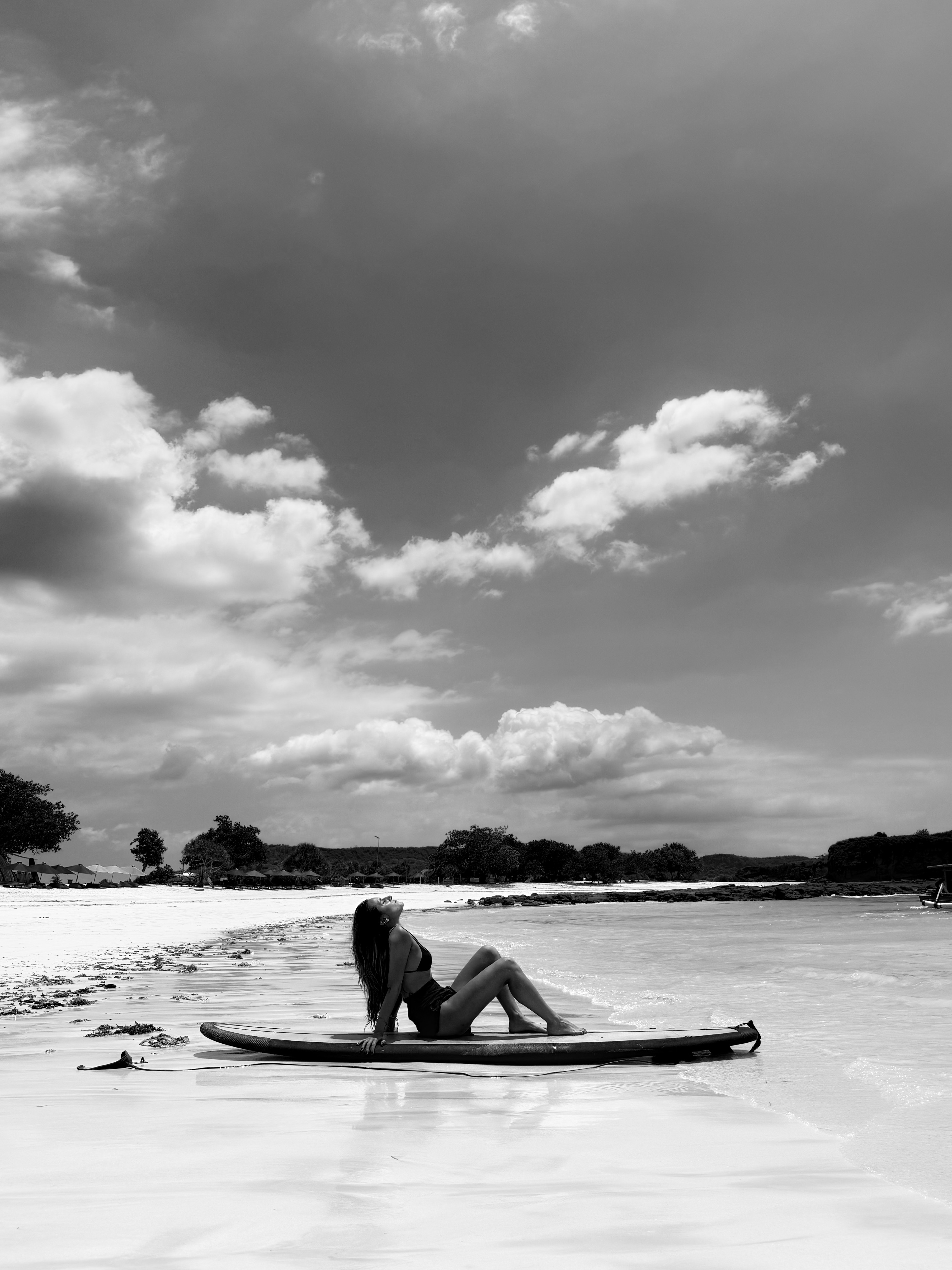 Woman in swimsuit sitting relaxed on a surfboard at the edge of a sandy beach under a cloudy sky.