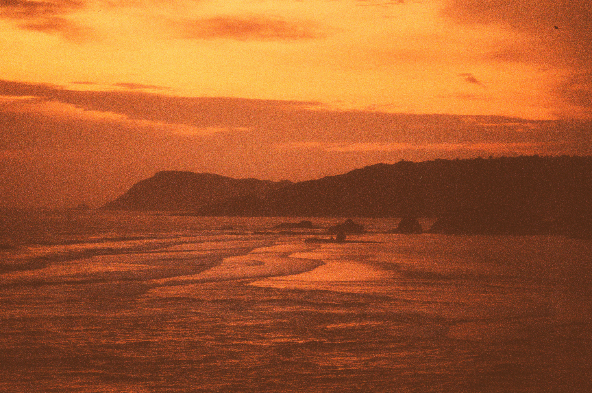Orange-tinted sunset over a calm ocean with a distant hilly coastline and gentle waves.