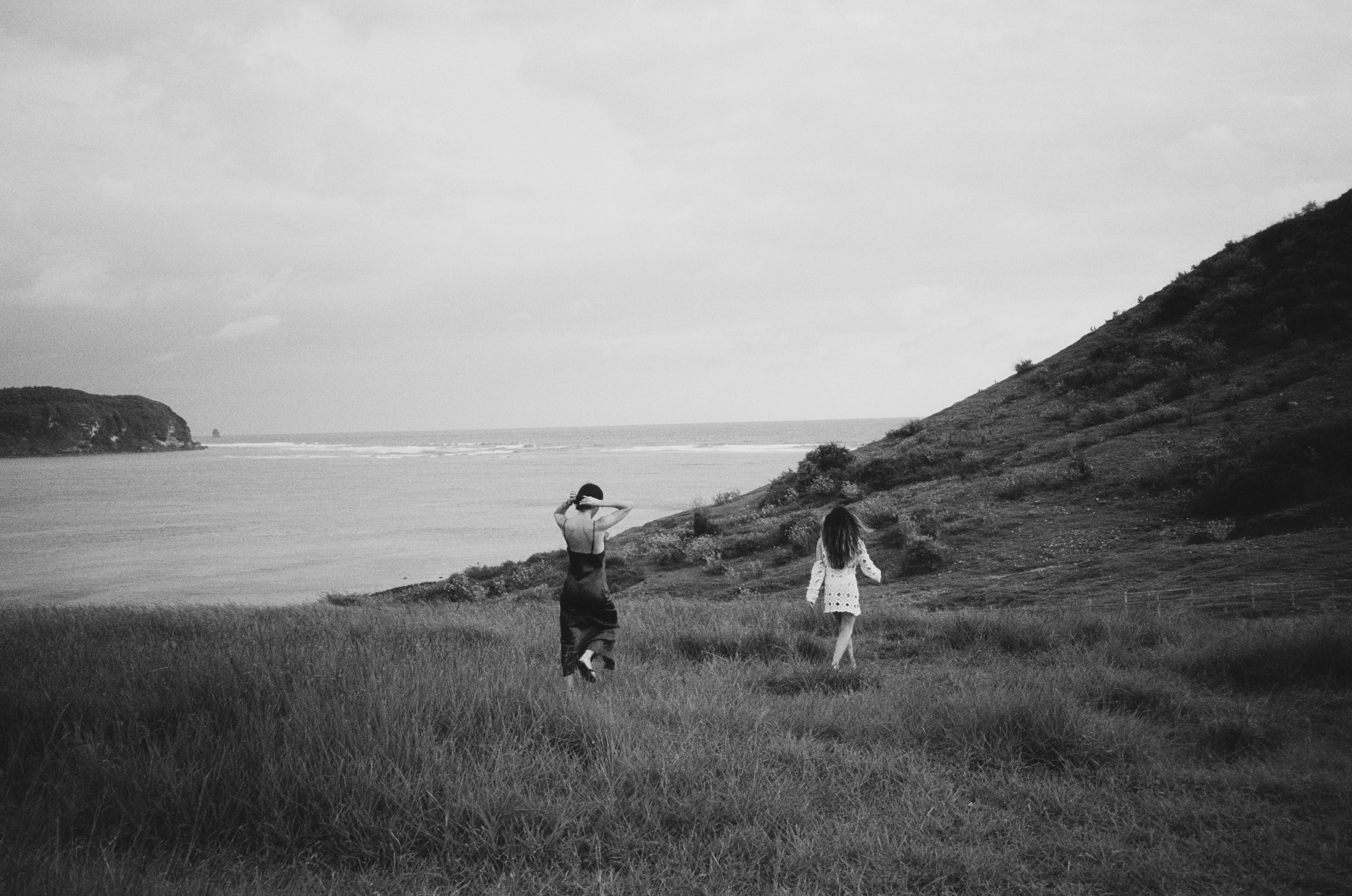 Two women walking through tall grass toward the ocean near a hillside on a cloudy day.