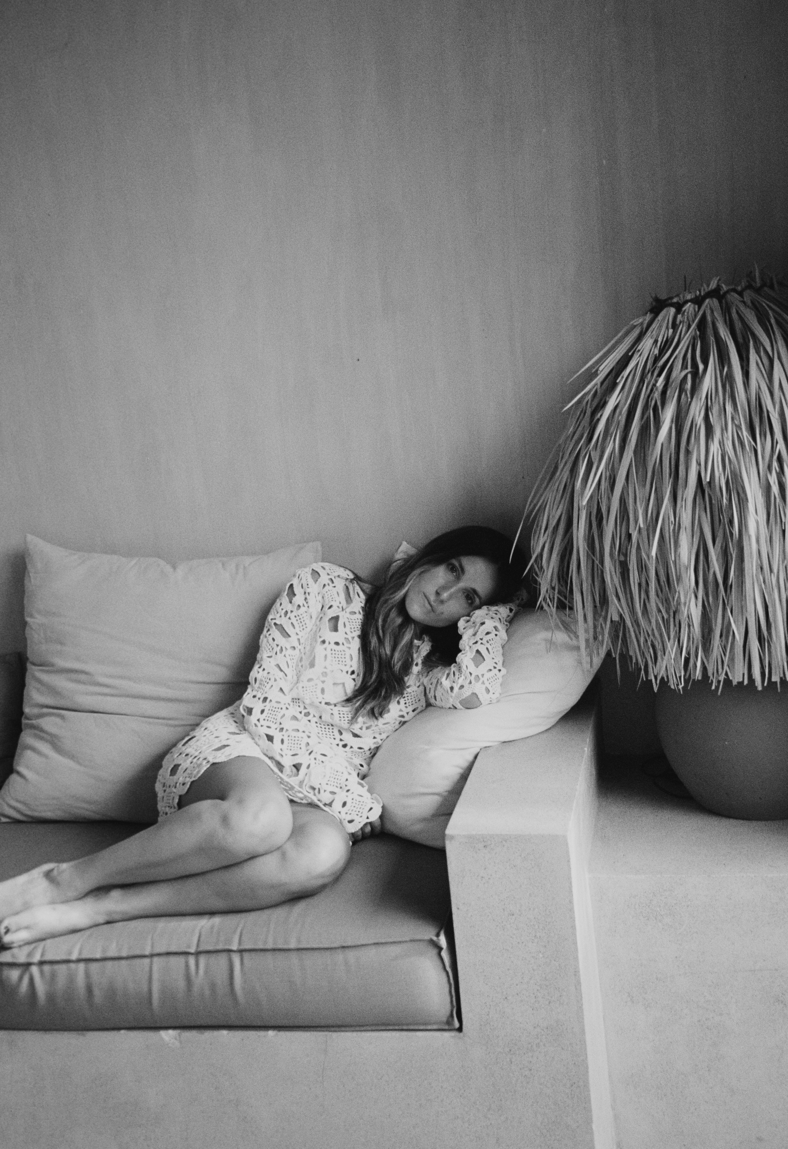 Woman in a white lace dress reclining on a cushioned bench next to a large potted plant with long leaves.