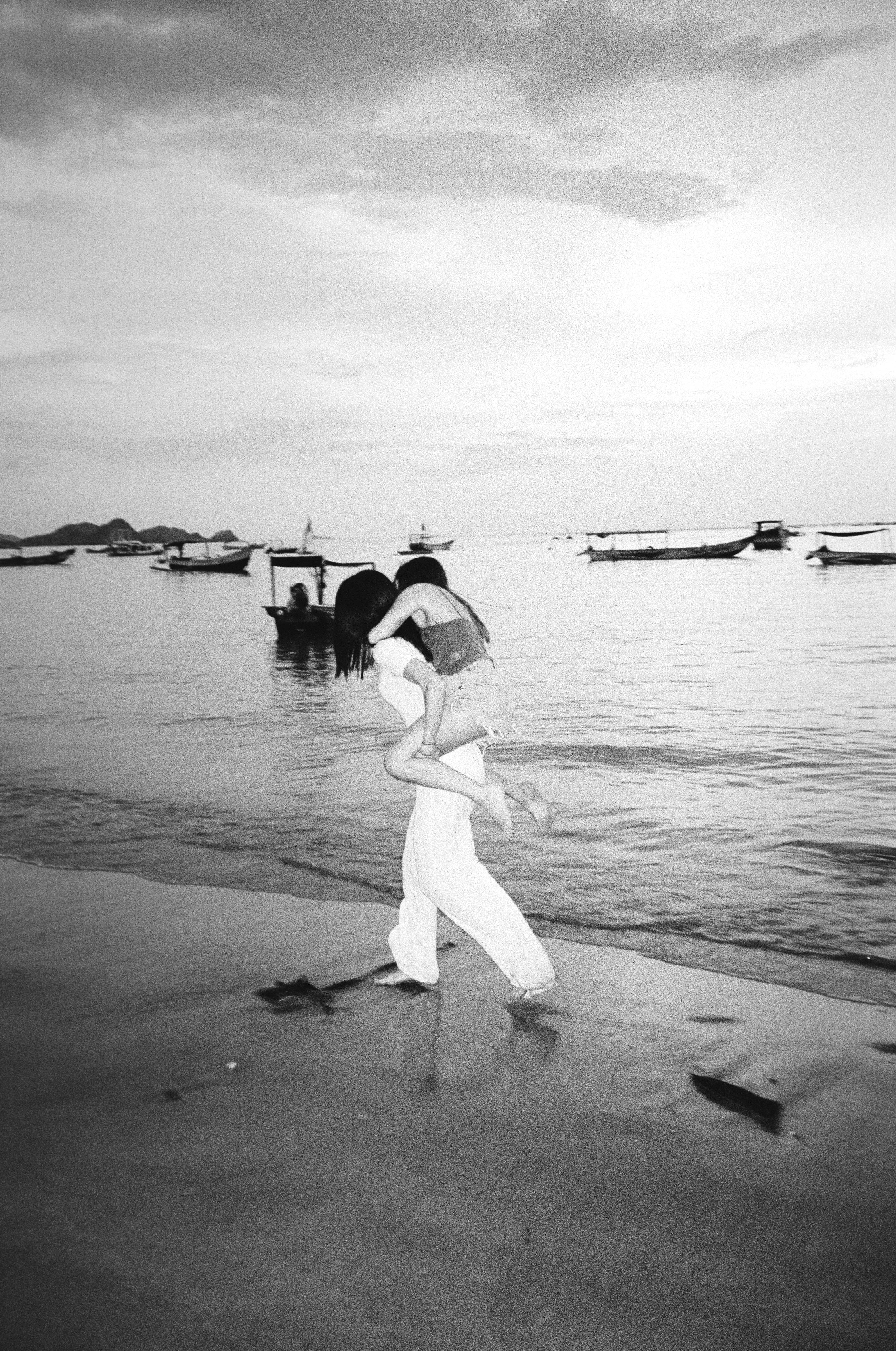 Black and white photo of a person giving a piggyback ride to another on a beach with several boats in the water.