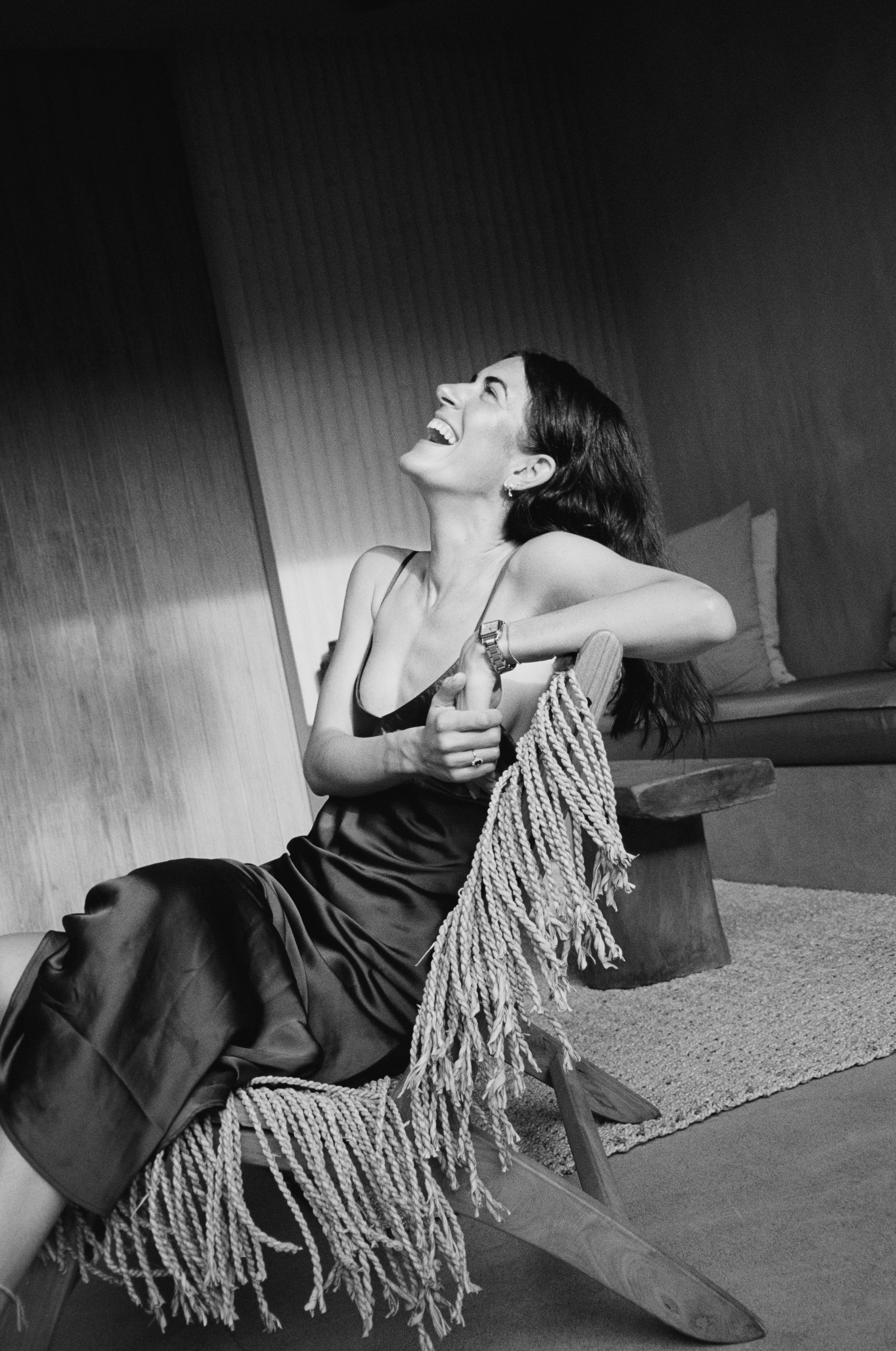 Smiling woman in a satin dress sitting on a fringed wooden chair indoors.